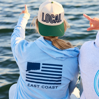 Person wearing a 'LOCAL' cap and 'EAST COAST' hoodie by a body of water. The Two Oh Three brand The 203