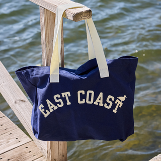 A navy blue canvas tote bag with cream handles and 'EAST COAST' printed in cream on the front, hanging from a wooden dock railing with water in the background.